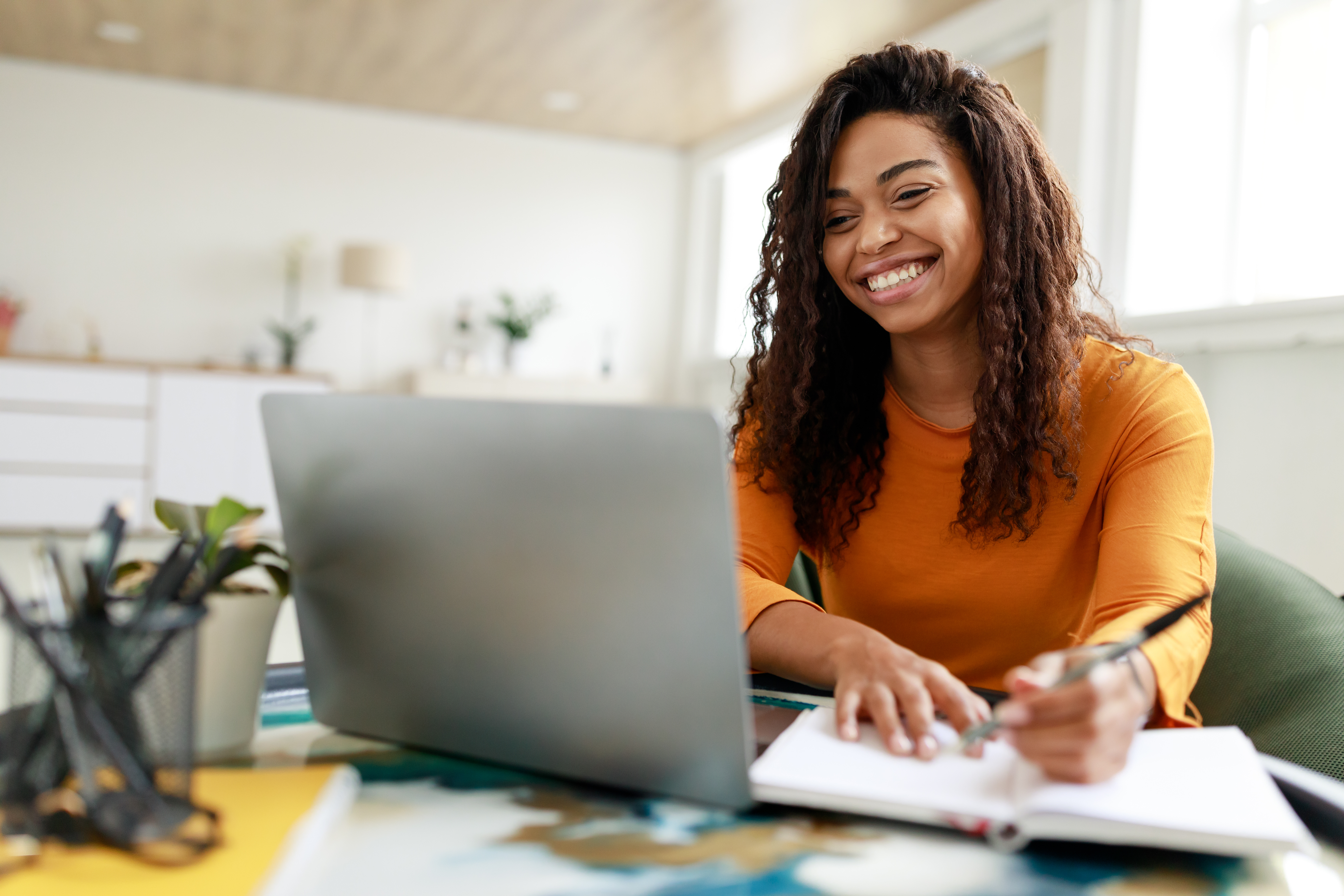 Woman looking at a laptop and smiling as she writes in a notebook