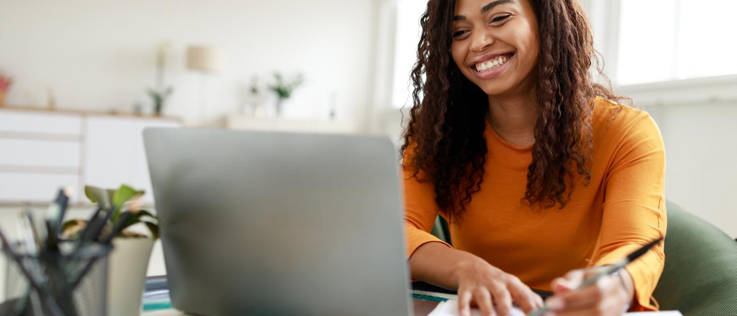 Woman looking at a laptop and smiling as she writes in a notebook